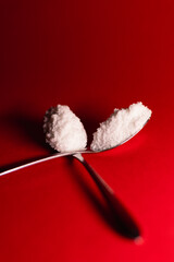 Close-up still-life of two metal spoons filled with white granulated powder on a vivid red background, creating a striking contrast with dramatic lighting and clean composition.