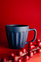 Festive still-life featuring a blue ceramic mug placed against a vibrant red background, accented with glossy red berries for a warm, seasonal holiday atmosphere.
