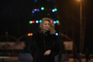 Woman with long blonde hair, dressed in a black fur coat, stands outdoors surrounded by colorful...