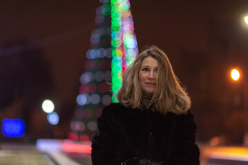 Woman with long blonde hair, dressed in a black fur coat, stands outdoors surrounded by colorful...