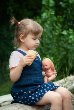 Little Girl Sitting Outdoors Holding a Toy Doll and Ice Cream Cone on a Summer Day