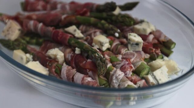 Green asparagus bundles in bacon with gorgonzola cubes set in a glass baking dish on white.