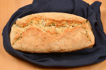 A freshly baked loaf of bread with a golden-brown crust and visible seeds rests on a dark blue waffle-weave cloth on a wooden surface