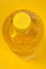 A high-angle close-up view of a clear plastic bottle with a yellow screw cap, filled with light yellow oil, set against a solid yellow background