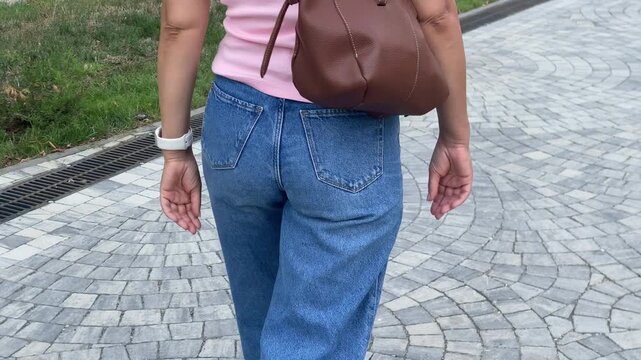 A casual woman walks away on a grey stone pavement. She wears blue jeans and a brown backpack while strolling past green grass during a summer day outdoors.