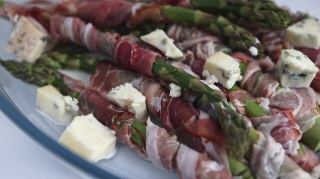 Bacon-wrapped asparagus arranged with gorgonzola cubes in a glass baking dish on white backdrop.