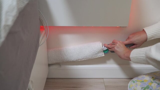 A meticulous woman uses a long microfiber duster to clean a tight gap behind furniture. She carefully removes dust from a hard-to-reach spot near the baseboard in a room with red LED lighting.