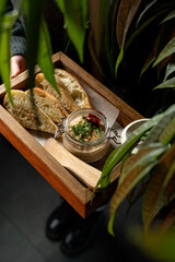 A person holds a wooden tray featuring artisanal bread and a small dish topped with herbs and a red chili. Lush green plants surround the cozy café scene, creating a warm atmosphere