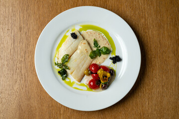 A beautifully arranged plate features tender white fish fillets drizzled with a creamy sauce, surrounded by fresh cherry tomatoes, herbs, and an artistic touch of caviar