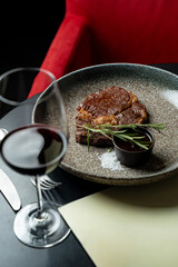 Juicy steak placed on a textured plate, garnished with rosemary sprigs, sits beside a glass of rich red wine in a chic restaurant. Elegant red chair adds sophistication