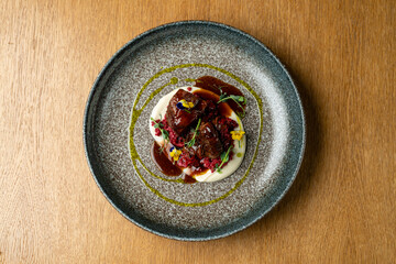 Tender meat sits atop a smooth and creamy puree, beautifully garnished with colorful herbs and edible flowers on a rustic wooden table. The culinary artistry shines in this elegant presentation