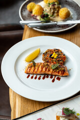 A beautifully presented dish features grilled salmon adorned with sauce and fresh herbs, accompanied by a colorful vegetable patty and a wedge of lemon. The elegant setting enhances the feast