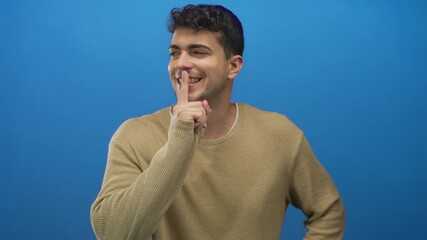 Man places finger to shush in studio as guy young hispanic smiling against blue background.