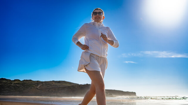 Beautiful middle-aged woman running along ocean on sandy Bordeira beach in Portugal on sunny summer day. Front view	