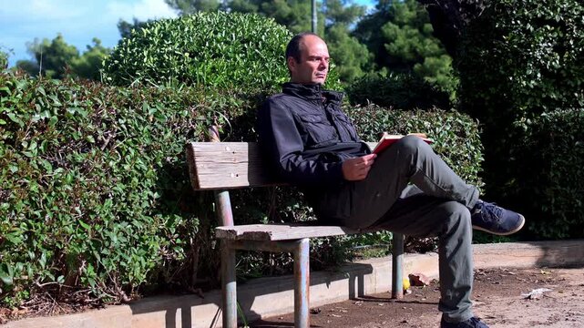 A  middle aged man is sitting on  a bench at a park alone and is reading a book on a sunny winter day.