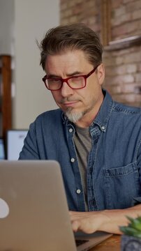 Happy older man working at home with laptop computer sitting at desk in home office. Zoom in.