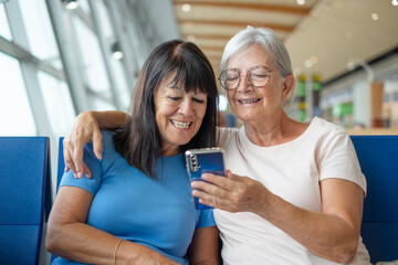 Smiling bonding couple of senior friends sharing social media on mobile phone sitting in airport departure area waiting for boarding. Travel and tourism concept, retiree lifestyle