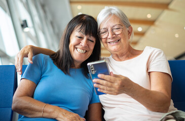 Cheerful bonding couple of senior friends sharing social media on mobile phone sitting in airport departure area waiting for boarding. Travel and tourism concept, retiree lifestyle