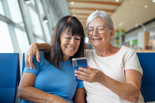 Smiling bonding couple of senior friends sharing social media on mobile phone sitting in airport departure area waiting for boarding. Travel and tourism concept, retiree lifestyle - Powered by Adobe