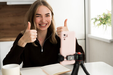 Portrait of happy Caucasian woman with thumbs-up during smartphone blogging looking at camera on her kitchen, shooting influence vlog
