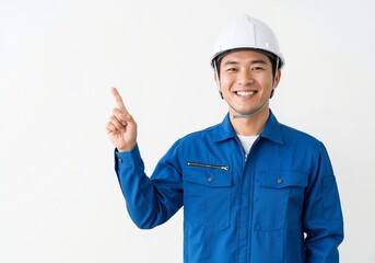Smiling asian engineer in a hard hat pointing upwards. Professional construction worker in a blue uniform presenting something on a white background with copy space