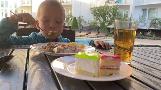 A young boy holds a fork while eating dinner at an outdoor table. He sits near a swimming pool with a plate of pasta and salad, accompanied by two pieces of cake and a drink.