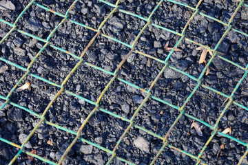 A textured close-up showcases the intersection of a green-blue wire mesh, creating a diagonal pattern in the foreground against a contrasting backdrop of dark stone fragments or asphalt.