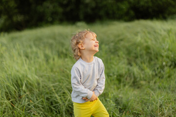 Smiling toddler standing in tall green grass on a sunny day. Candid outdoor moment with warm light, joyful expression and natural childhood energy.