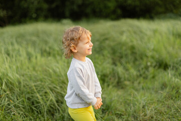 Smiling toddler standing in tall green grass on a sunny day. Candid outdoor moment with warm light, joyful expression and natural childhood energy.