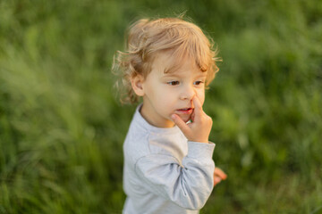 Curious toddler touching his nose while standing outdoors on a green field. Natural candid moment reflecting real childhood behavior in soft evening light.