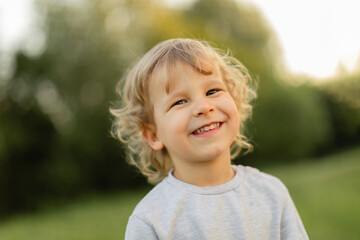 Smiling blonde toddler outdoors in soft natural light, captured with a warm candid expression against a blurred green background. Happy childhood moment.