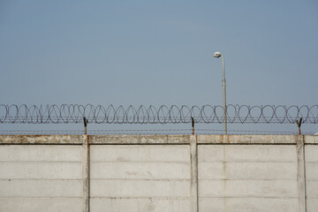 Fototapeta premium Concrete security wall with razor wire fence and street light pole against blue sky background.