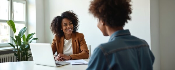 Smiling Black businesswoman consults female client at office desk. Discusses finance, investment plans with customer, offering advice, guidance on future goals. Two women meet with pro advisor.