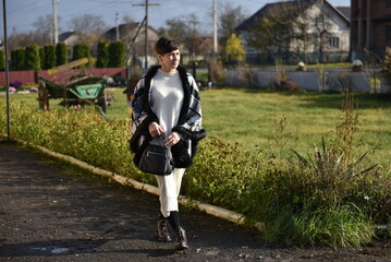 Obraz premium Woman standing outdoors on a sunny day, looking to the side while holding a small black bag, dressed in a warm light sweater and plaid coat