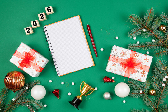 Planning New Year 2026 celebrations. Notepad for wishes and gift ideas, surrounded by festive decorations. Top view on Christmas green background with ample copy space for text.