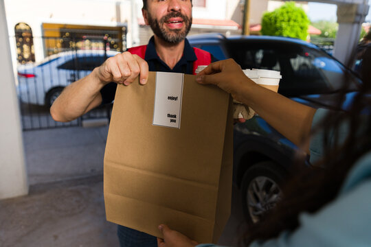Closeup of delivery man handing a paper bag with food and coffee cups to a young woman receiving the order at home
