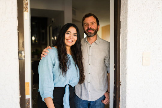 Couple welcoming guests at front door, smiling happy at apartment entrance, husband and wife standing together at home - Powered by Adobe
