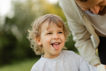 Happy toddler sticking out tongue while playing outdoors, with parent leaning in beside him. Warm natural light, candid family moment full of joy and connection.