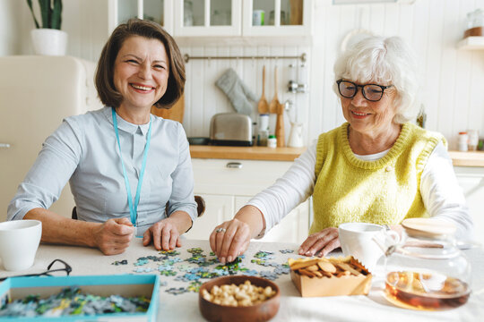 Two cheerful ladies playing puzzle together at kitchen table, mature female therapist working with senior patient with dementia using method of memory stimulation through board games - Powered by Adobe
