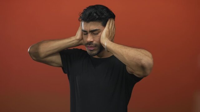 Young hispanic man covers ears with hands against orange studio wall; sound sensitivity overload anxiety.