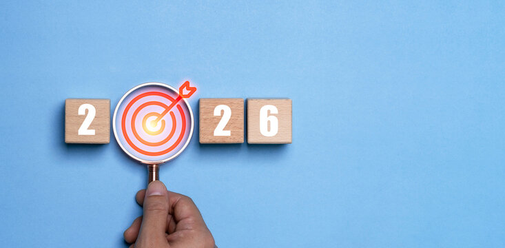 Hand holding magnifying glass highlighting a target icon between the numbers 2026 represents a happy new year with focus goal setting motivation precision planning and direction for future success.