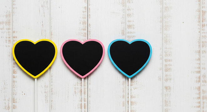 Three heart shaped chalkboard signs with yellow, pink, and blue border resting on a light rustic white wooden plank background for copy space