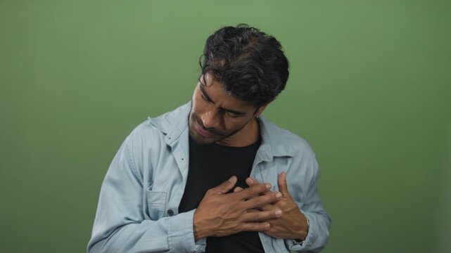 Man clutching chest in studio shot with serious worried expression against green background; pain distress.