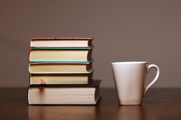 A large stack of books, on a wooden table, a white cup on the side