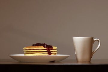 A white plate with pancakes drizzled with raspberry jam, a white cup on the side, all of this is on a brown wooden table, straight front view