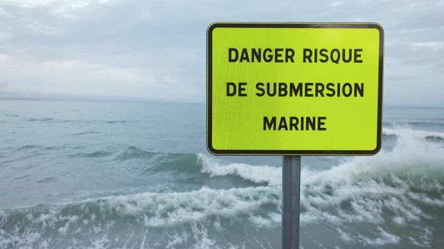 Slow motion turquoise sea storm waves crashing against a seawall, spraying foam around a danger marine submersion sign.
