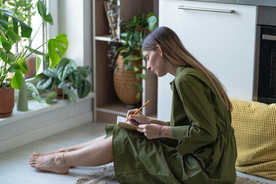 Relaxed middle aged woman with calm expression on floor in kitchen, writing thoughts ideas and gratitude list in notebook. Positive female taking notes journaling about feelings, mental health care.