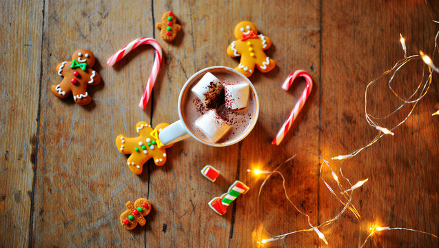 Cozy christmas morning scene with hot chocolate marshmallows gingerbread cookies candy canes and warm fairy lights on rustic wooden background overhead view Christmas Day - Powered by Adobe