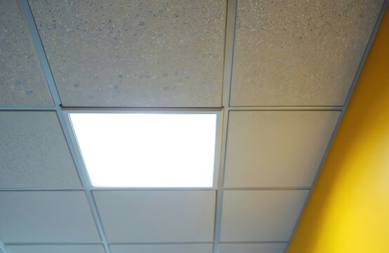 Modern suspended ceiling grid with bright square led light panel. Textured panels form geometric pattern above bright yellow wall. Interior architecture detail.