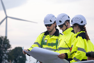 Workers examine plans for a wind farm project while wearing safety gear in a rural location during daylight hours